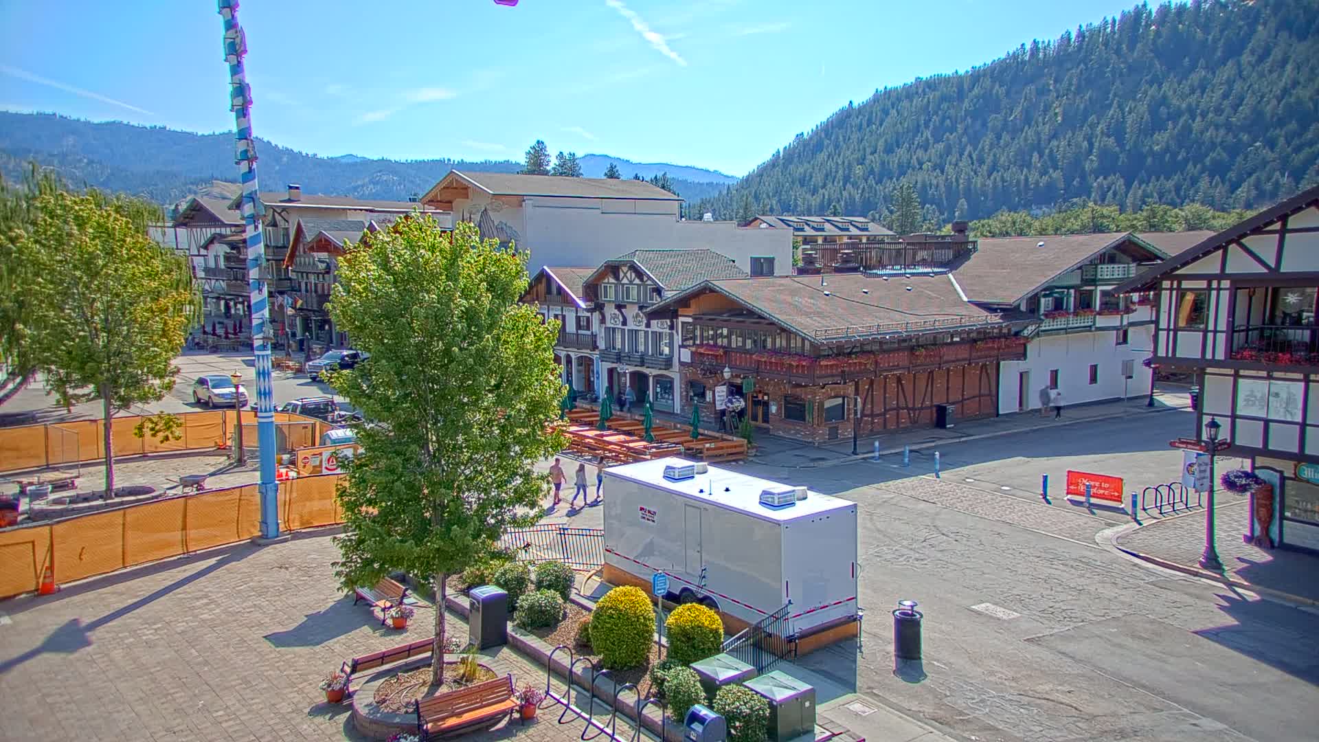 A town square with Bavarian-style buildings, a tall maypole, and a few people walking around on a sunny day, set against a backdrop of forested hills.