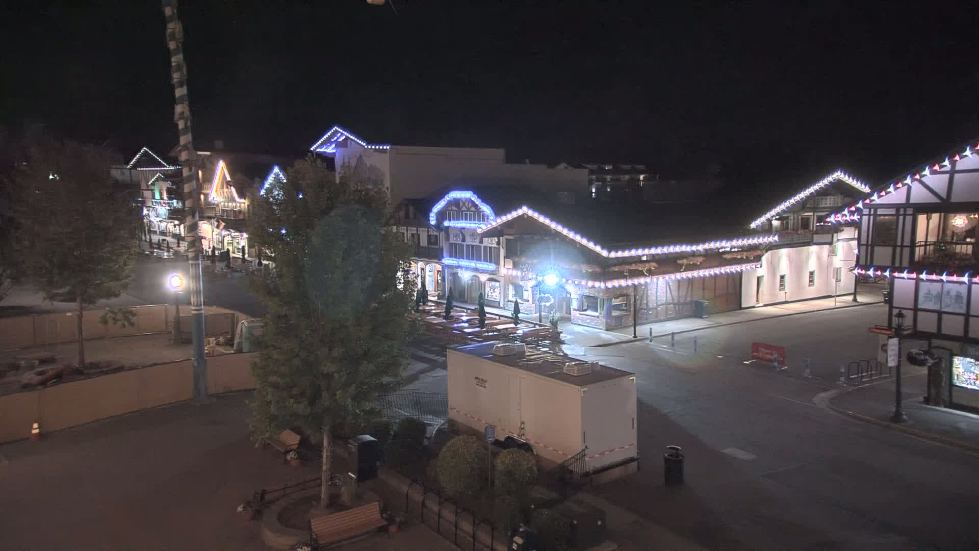 A nighttime, outdoor view of a street lined with illuminated buildings, some with festive lighting, and a large, empty space in the foreground.