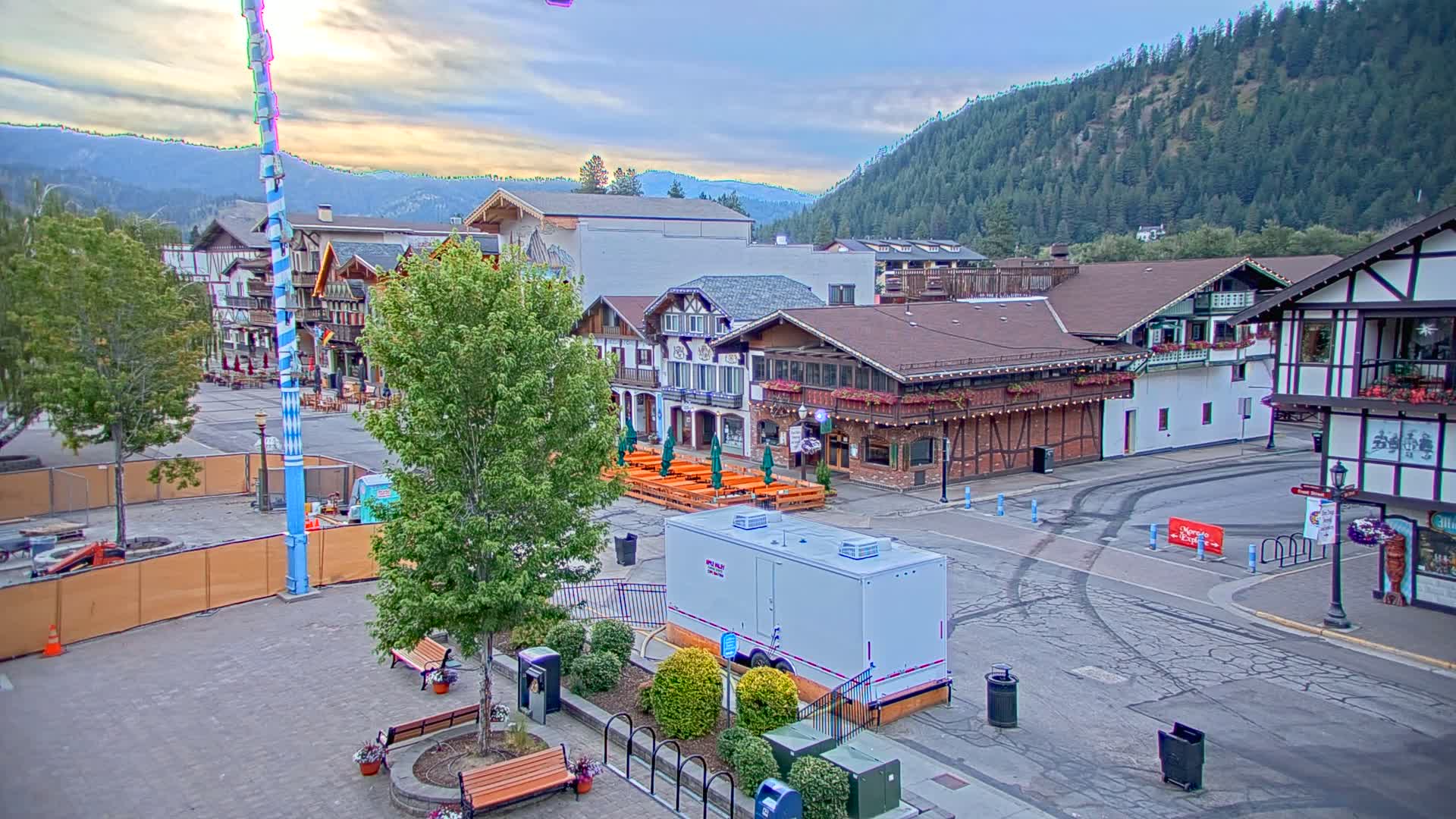 A town square with Bavarian-style buildings, outdoor seating, and a tall, decorated pole under a partly cloudy sky, backed by a mountain range.