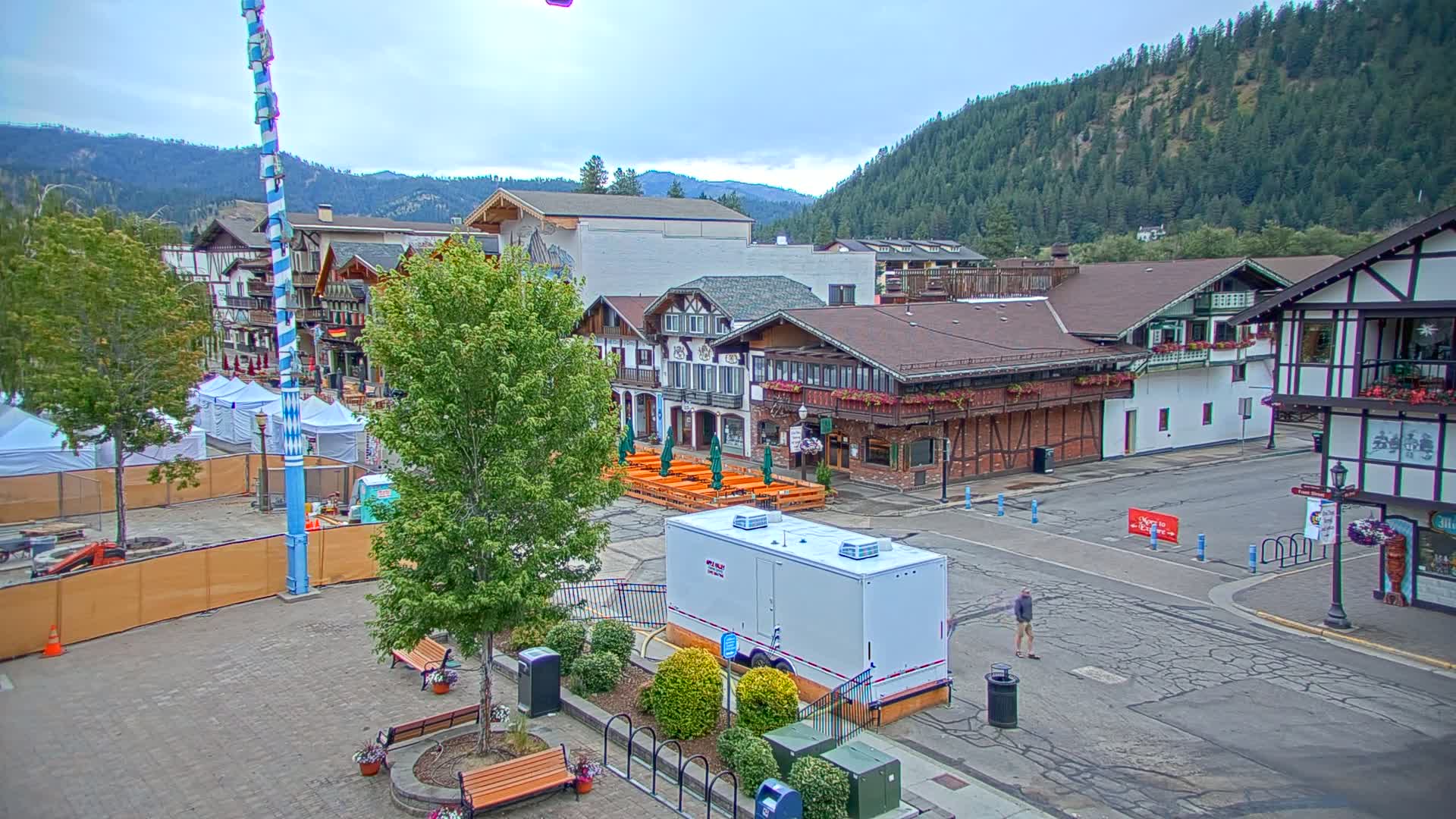 A town square with several buildings in a Bavarian style, surrounded by trees and mountains under an overcast sky.