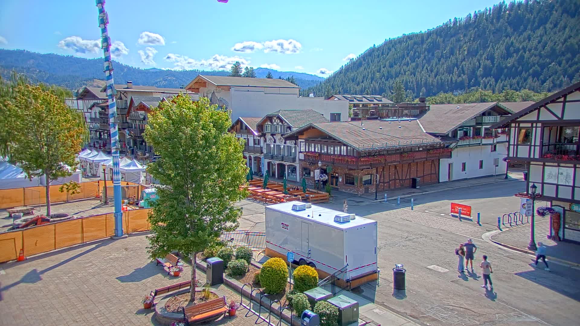 A town square with Bavarian-style buildings, a tall maypole, and several people walking on a sunny day with partly cloudy skies, nestled against a mountain backdrop.