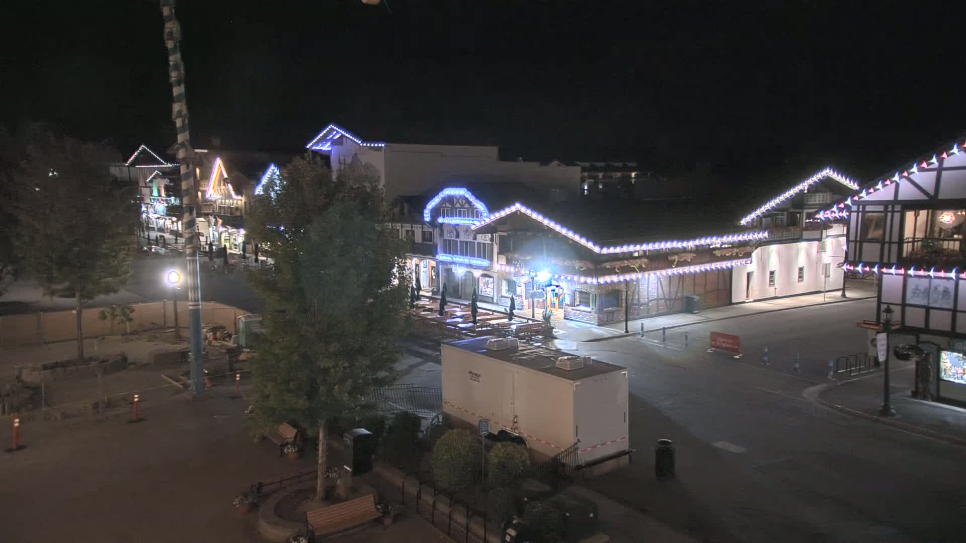 A nighttime, outdoor view of a town square with several buildings decorated with string lights, a portable restroom, and trees.