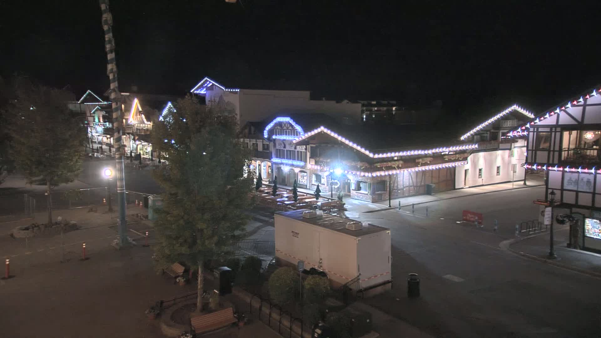A nighttime, street-level view of a row of festively-lit buildings, mostly empty, with a few trees and a portable restroom visible in the foreground.