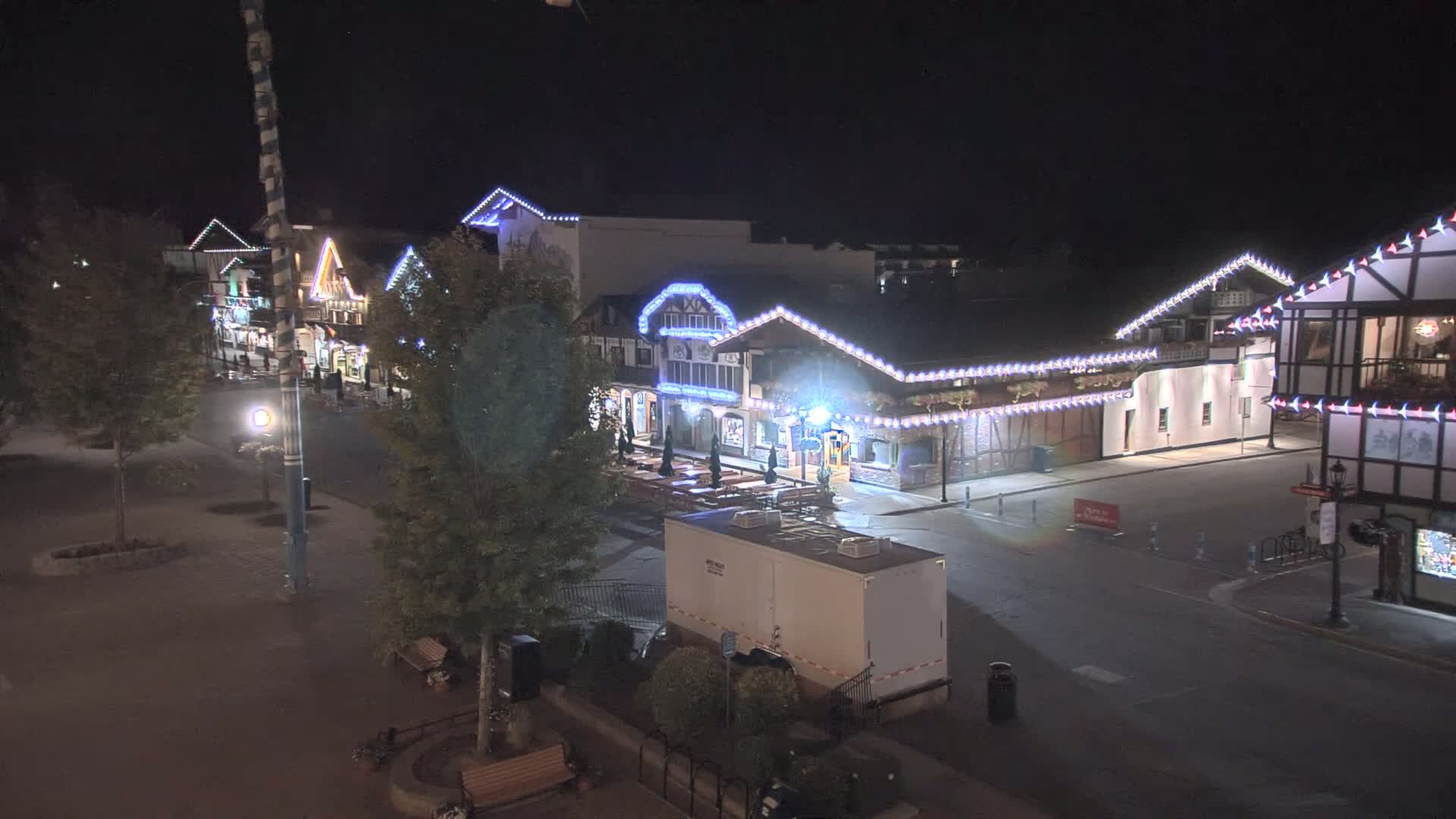 A nighttime view of a street lined with illuminated buildings, featuring string lights and trees.