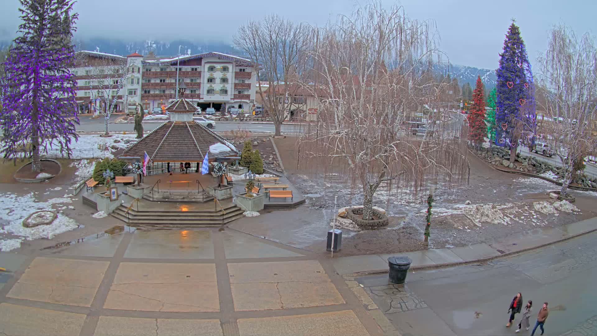 A town square with a gazebo, snow-covered ground, and several decorated Christmas trees under an overcast sky.