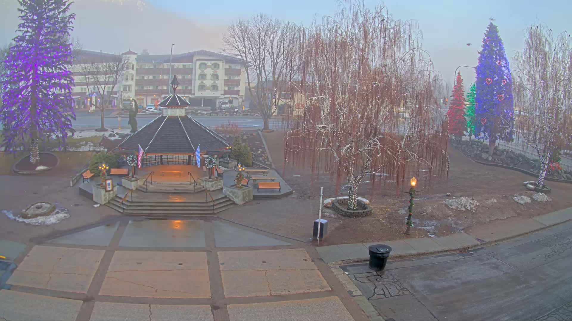 A gazebo with lights sits in a town square adorned with decorated trees and a building in the background, all under a foggy sky.