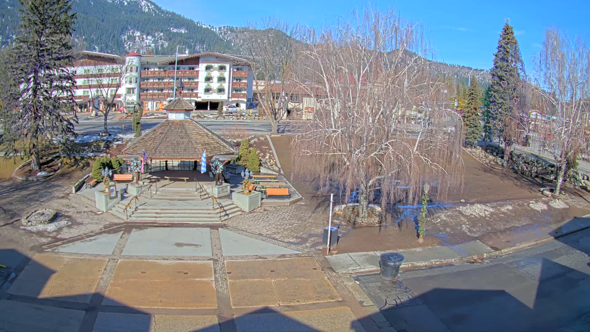 A sunny day reveals a town square with a gazebo, benches, and bare trees, partially muddy from melting snow, in front of a multi-story building and mountains in the background.