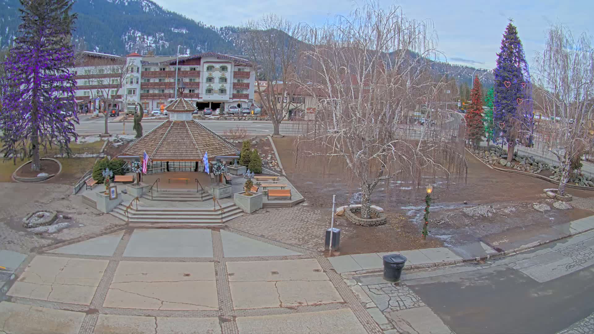 A town square with a gazebo, decorated trees, and a multi-story building in the background under a cloudy sky.