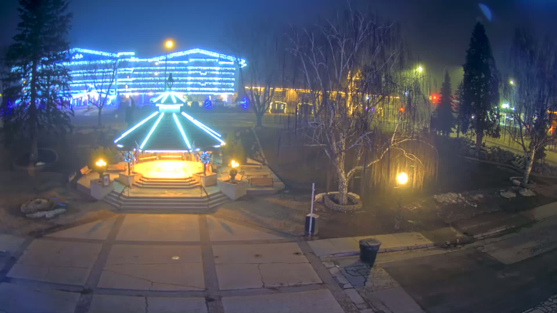 A brightly lit gazebo stands in a park at night, surrounded by bare trees and buildings decorated with Christmas lights under hazy conditions.