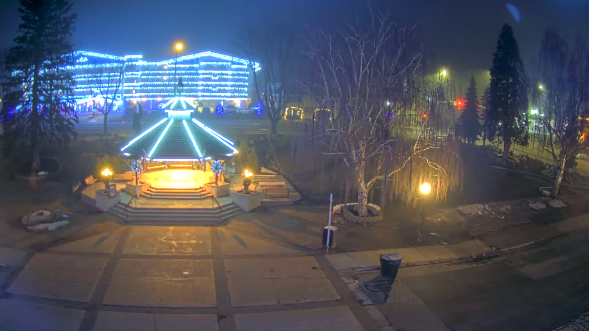 A brightly lit gazebo stands in a park at night, surrounded by bare trees and a building adorned with twinkling lights in a foggy atmosphere.
