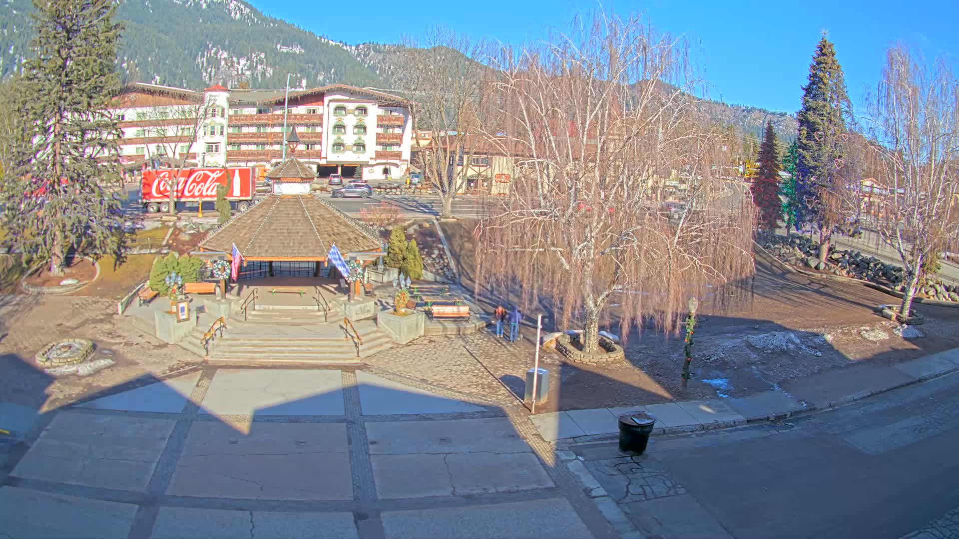 A plaza with a gazebo and a multi-story building in the background, on a sunny day with bare trees and snow-covered mountains in the distance.