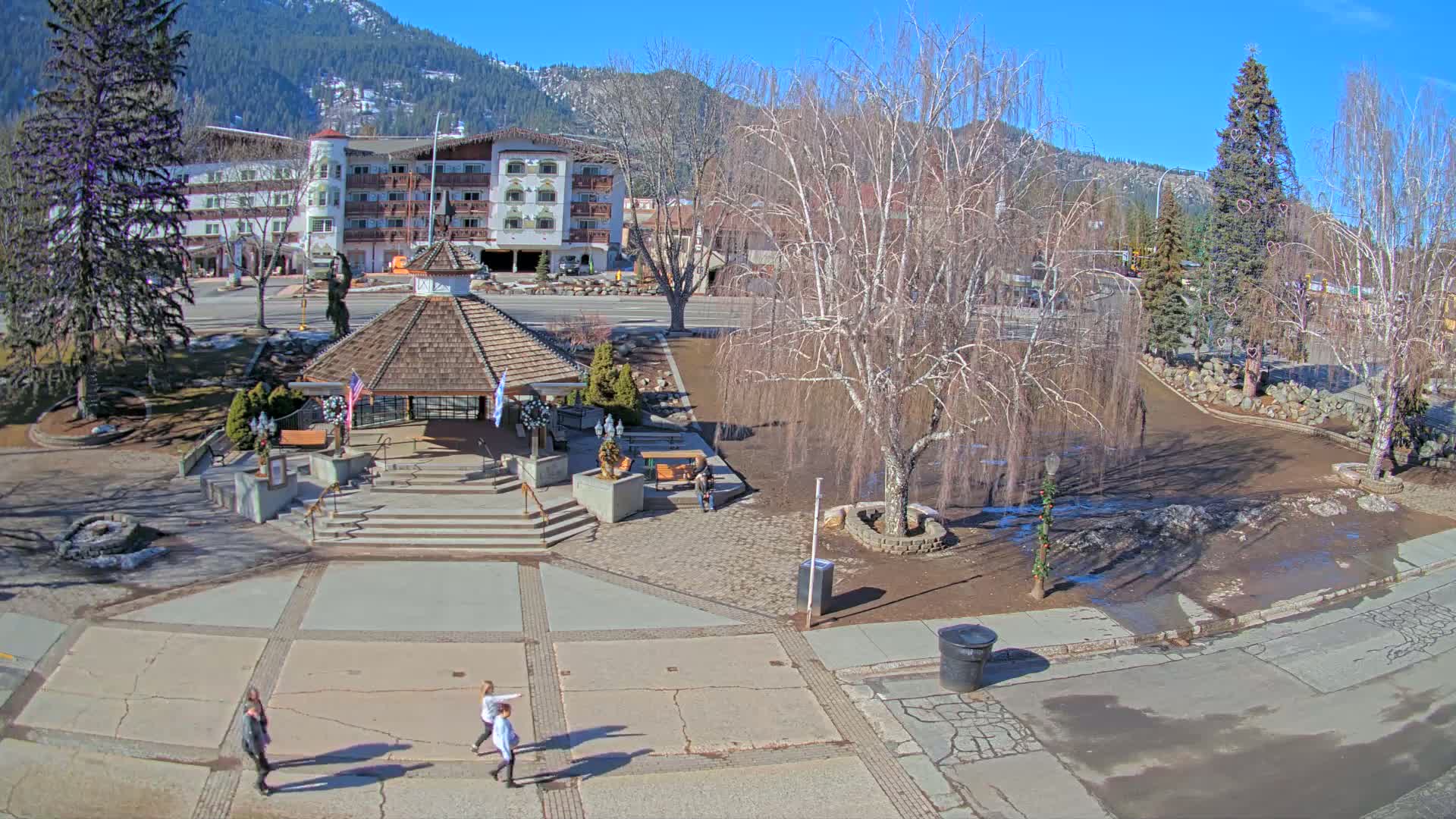 A sunny day reveals a town square with a gazebo, a multi-story building in the background, and several people walking on the pavement, all under a clear blue sky.