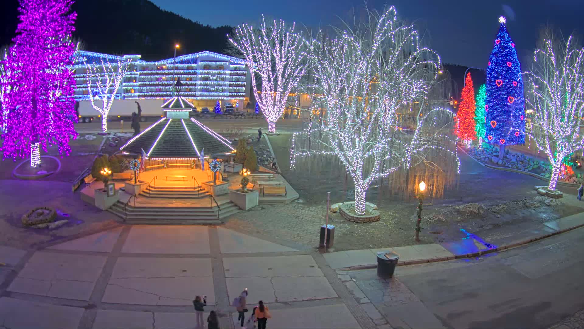 A town square at night is decorated with brightly lit Christmas trees and string lights on trees and buildings, with a gazebo in the center.