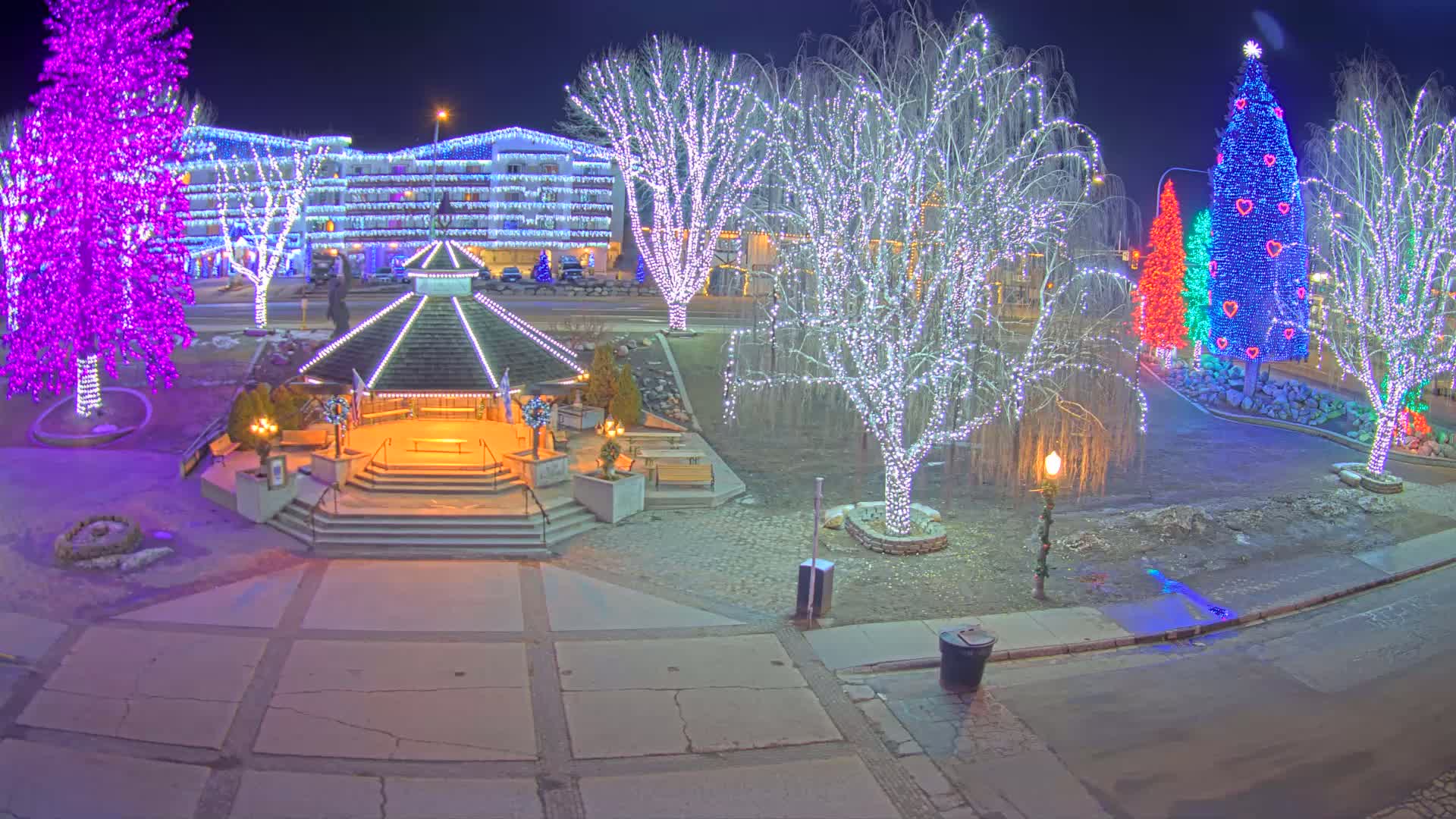 A nighttime scene of a town square decorated with colorful lights on trees and a gazebo, illuminated by soft lighting.