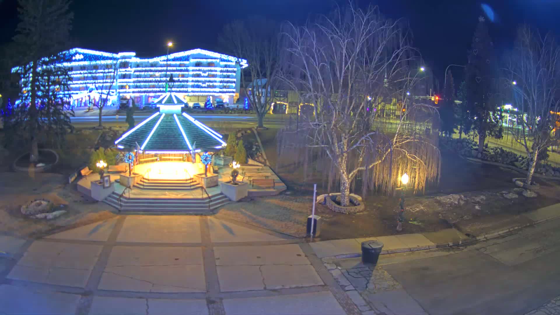 A brightly lit gazebo stands in a park at night, across the street from a building covered in Christmas lights.