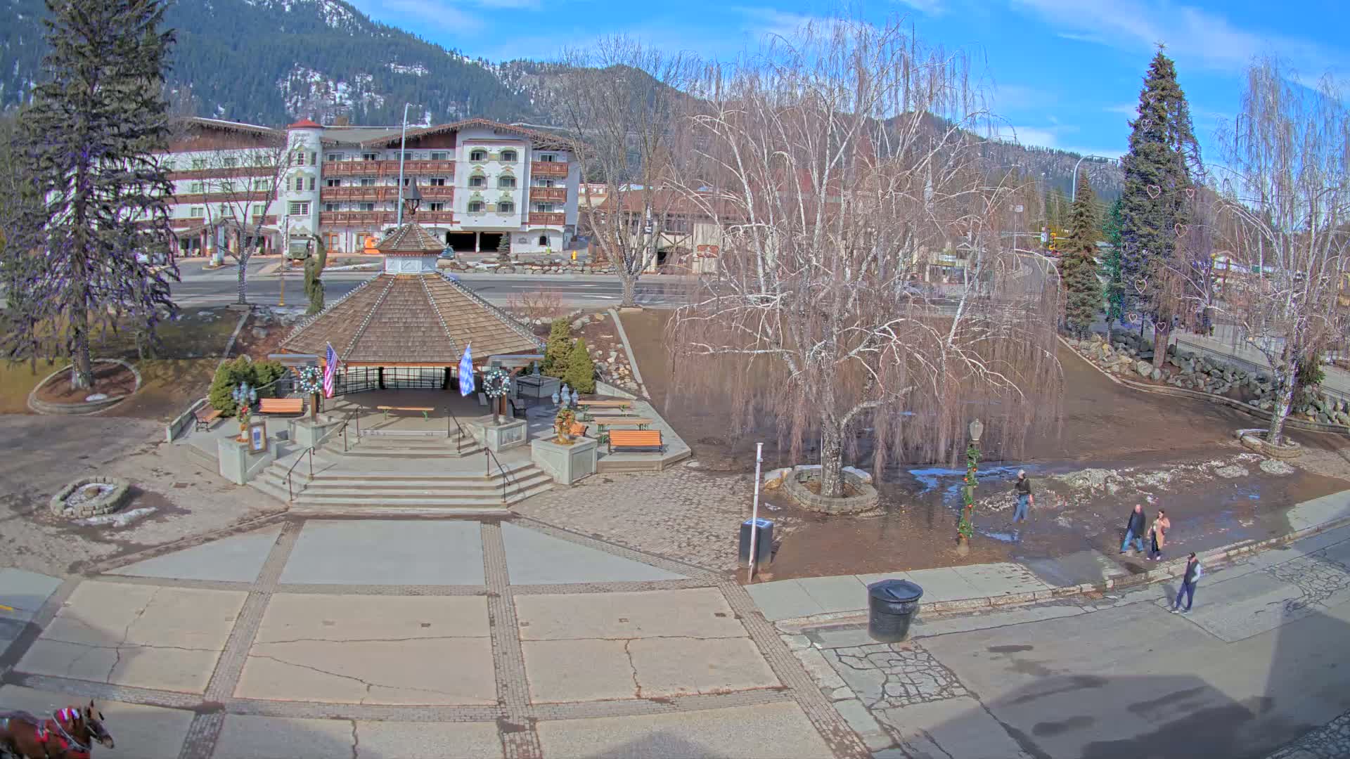 A town square with a gazebo, benches, and a large bare tree is partially muddy under a sunny sky, with a hotel and mountains in the background.