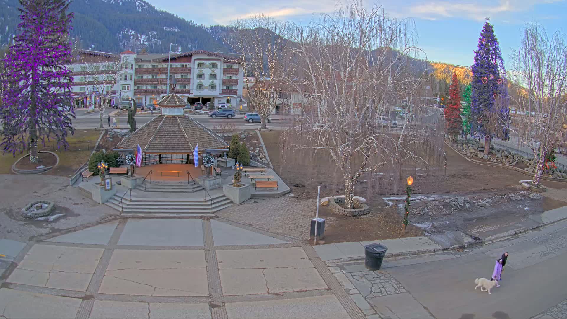 A town square with a gazebo, decorated trees, and a person walking a dog, all under a twilight sky.