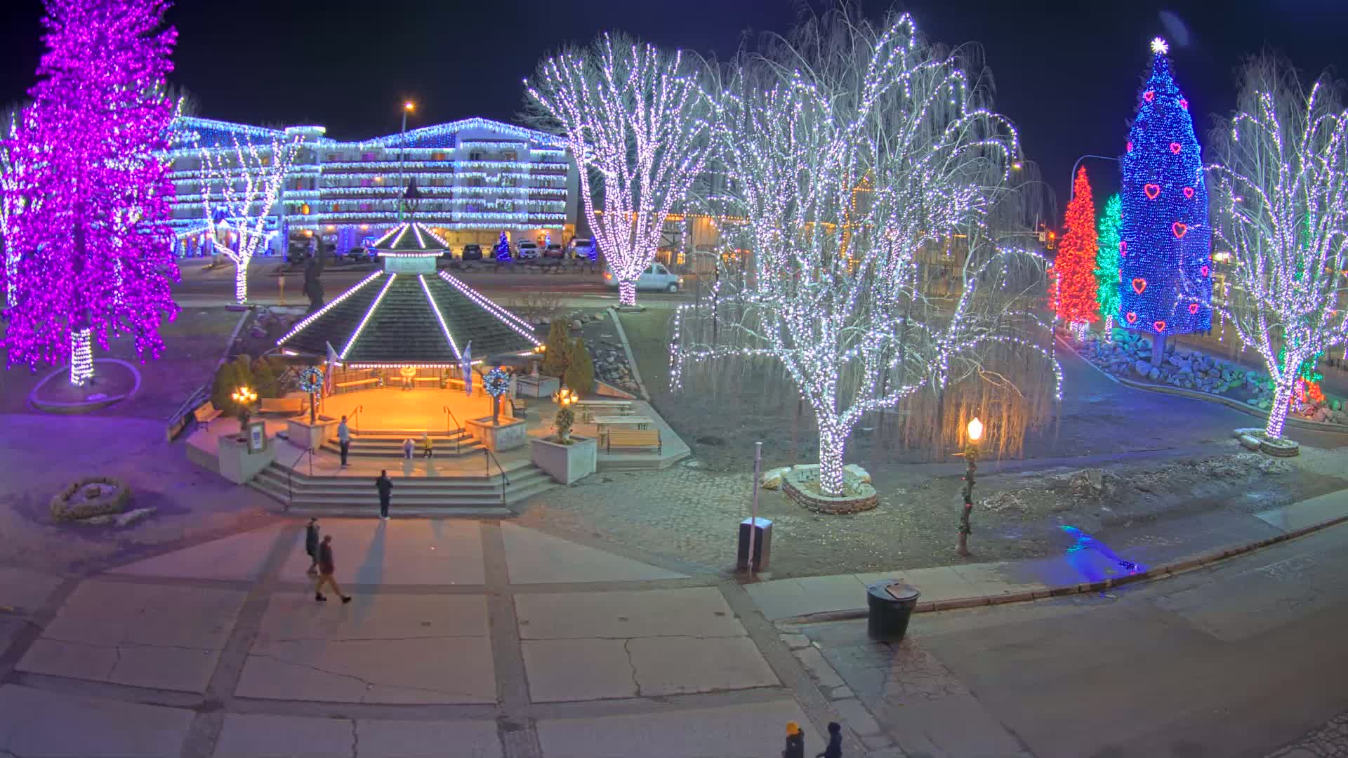 A nighttime scene of a town square decorated with colorful Christmas lights illuminating trees, a gazebo, and a building, with a few people walking around on a paved area.