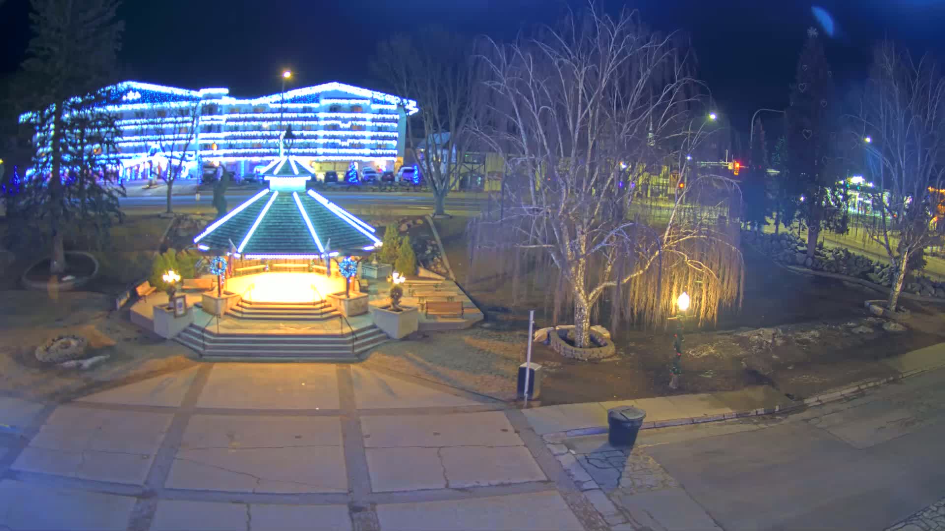 A brightly lit gazebo sits in a town square at night, next to bare trees and a building decorated with blue Christmas lights.