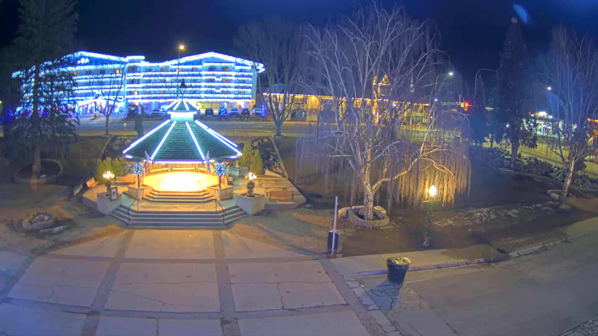 A brightly lit gazebo sits in a town square at night, with a large, illuminated hotel in the background and bare trees surrounding the area.