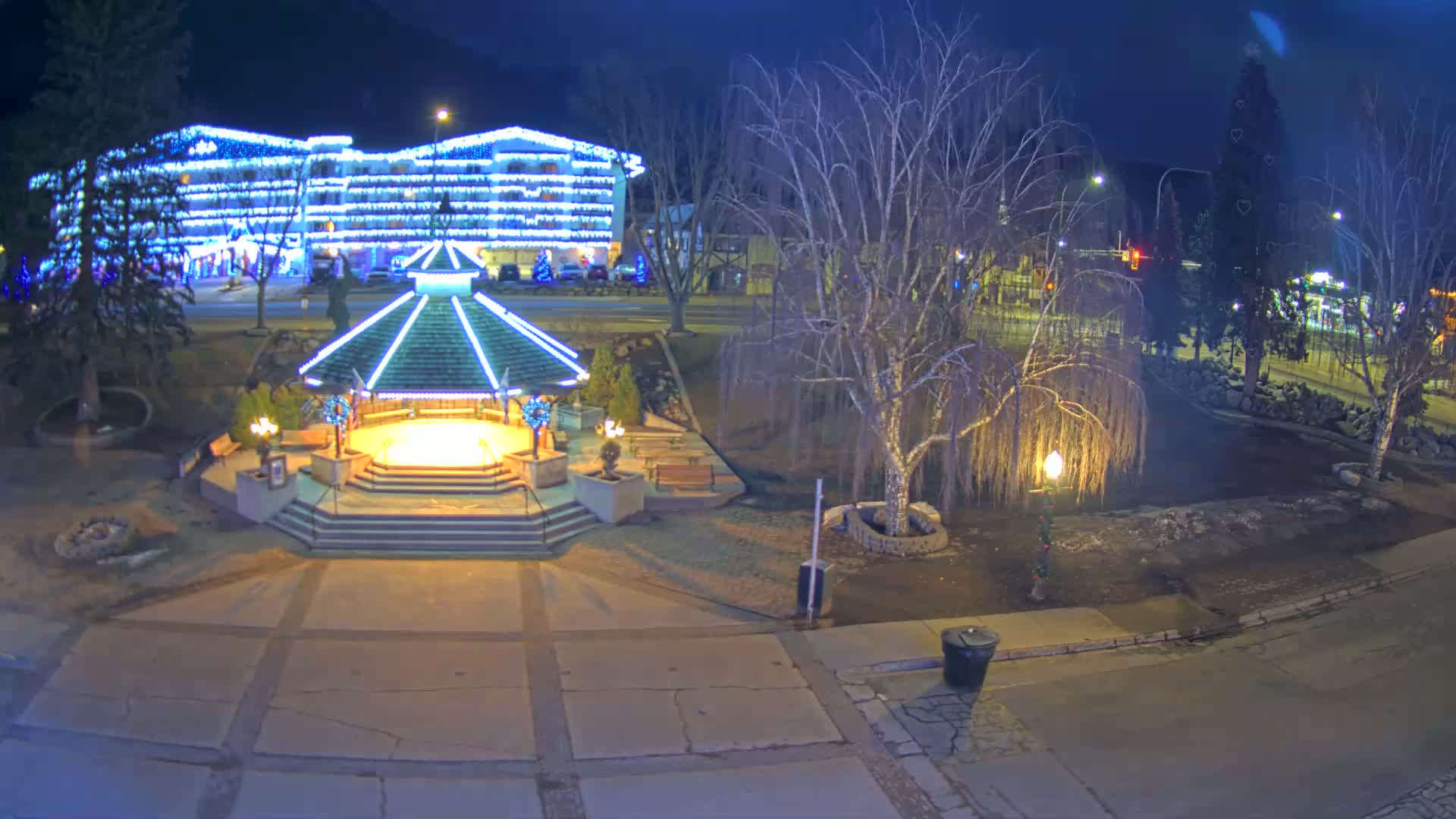 A brightly lit gazebo stands in a park at night, next to a building decorated with Christmas lights under a dark, clear sky.