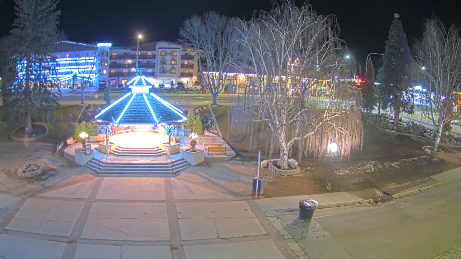 A brightly lit gazebo stands in a park at night, surrounded by bare trees and a snow-dusted town square, with a multi-story building decorated with Christmas lights in the background.
