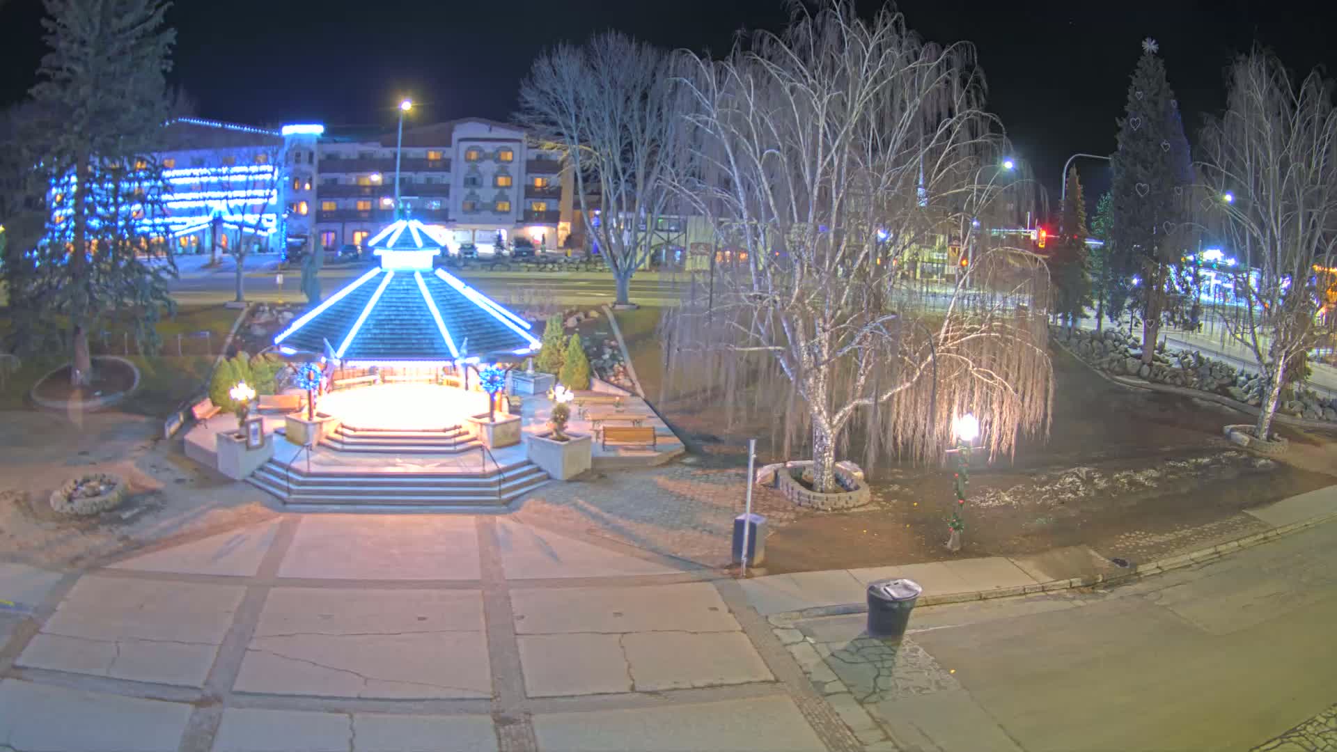 A brightly lit gazebo stands in a town square at night, surrounded by bare trees and a decorated Christmas tree, under a clear sky.