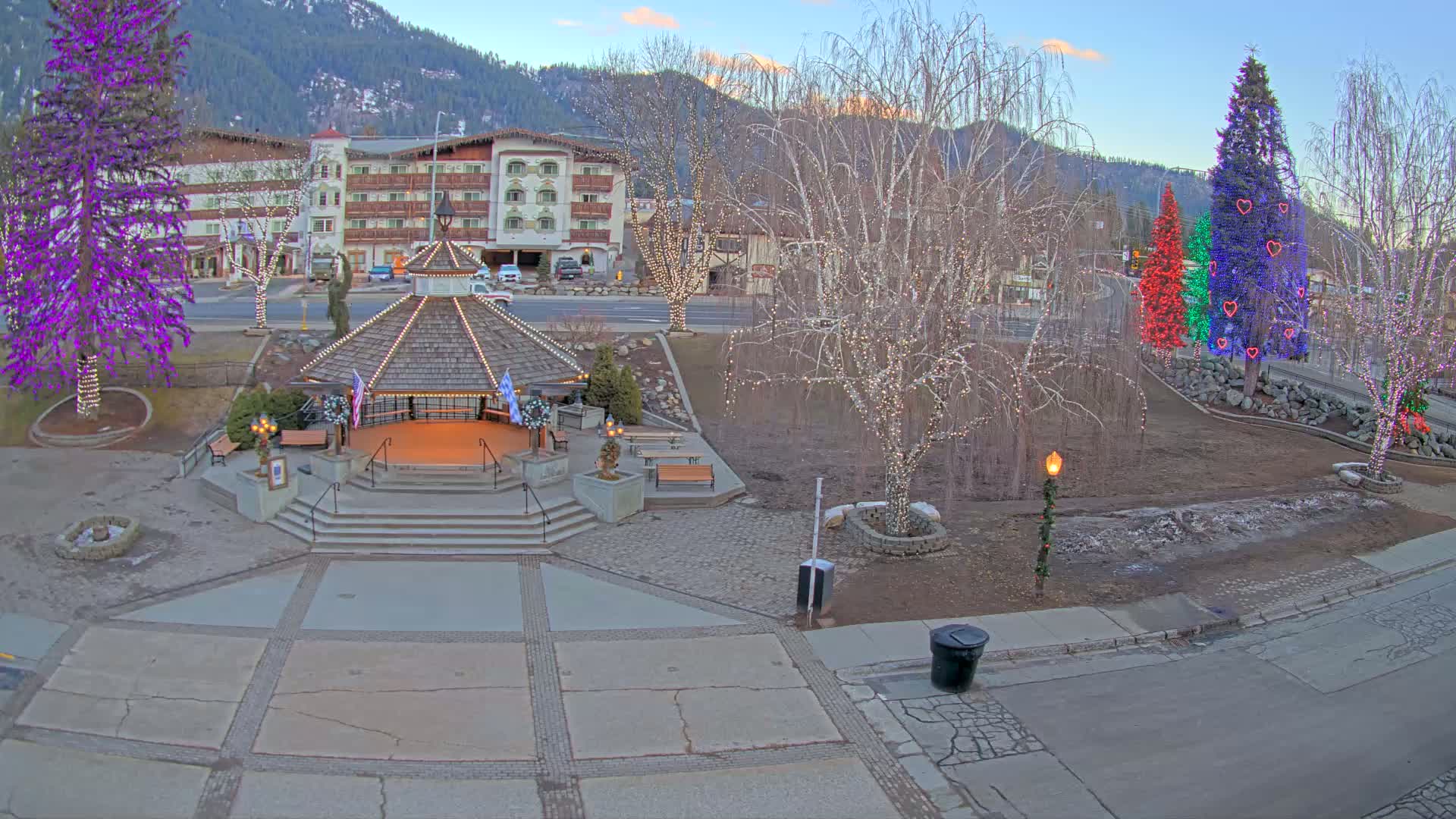 A town square with a gazebo, decorated trees, and a hotel in the background under a twilight sky.