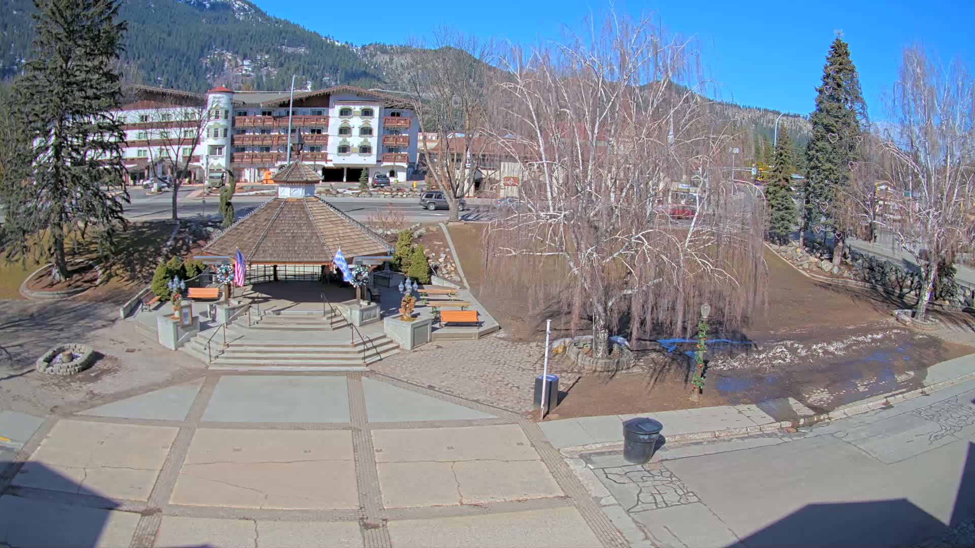A paved town square with a gazebo, benches, and bare trees under a sunny sky, in the background is a multi-story building and snow-capped mountains.