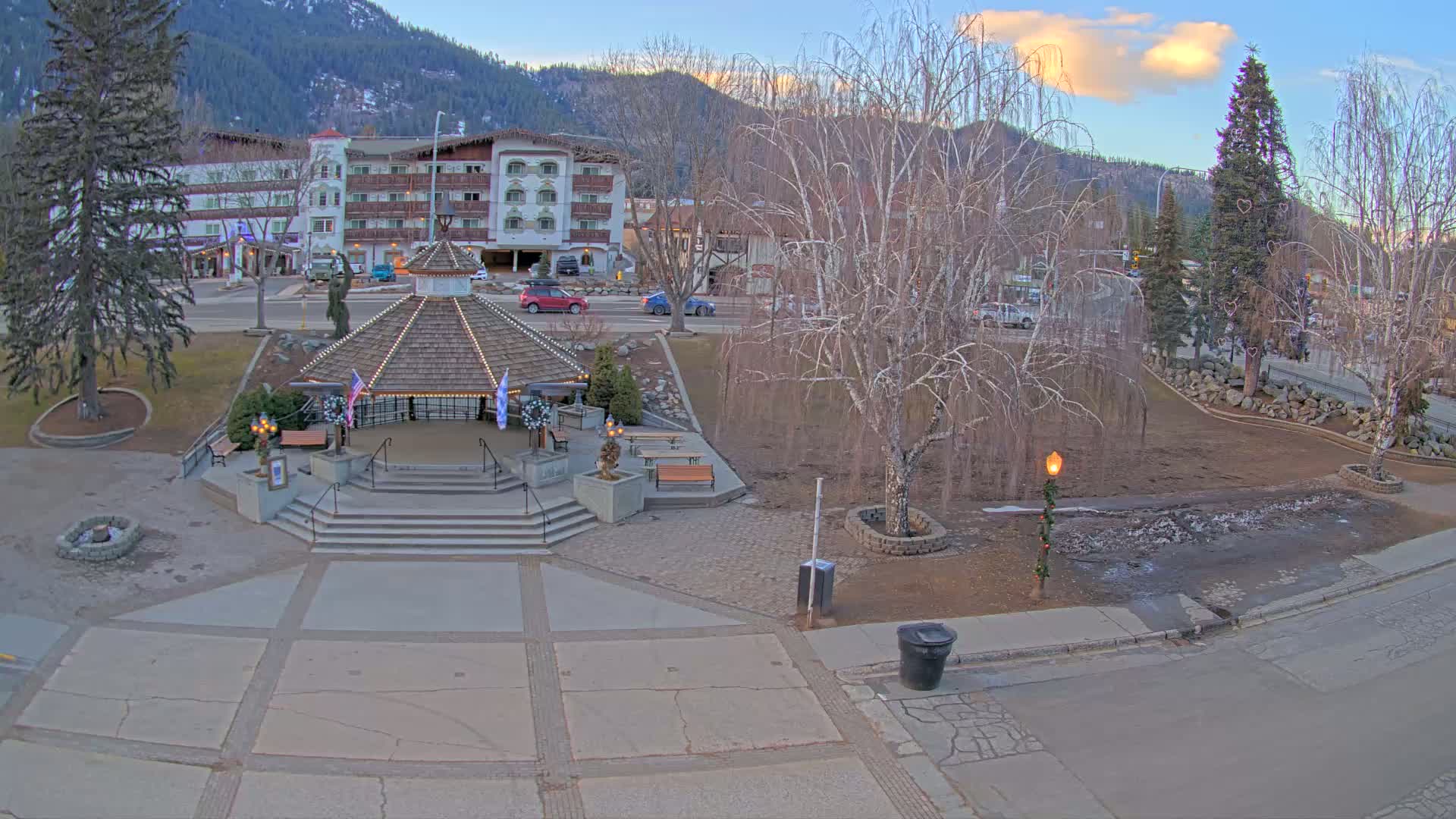 A gazebo sits in a town square, next to a partially melted patch of snow, with bare trees and a hotel visible in the background under a partly cloudy evening sky.