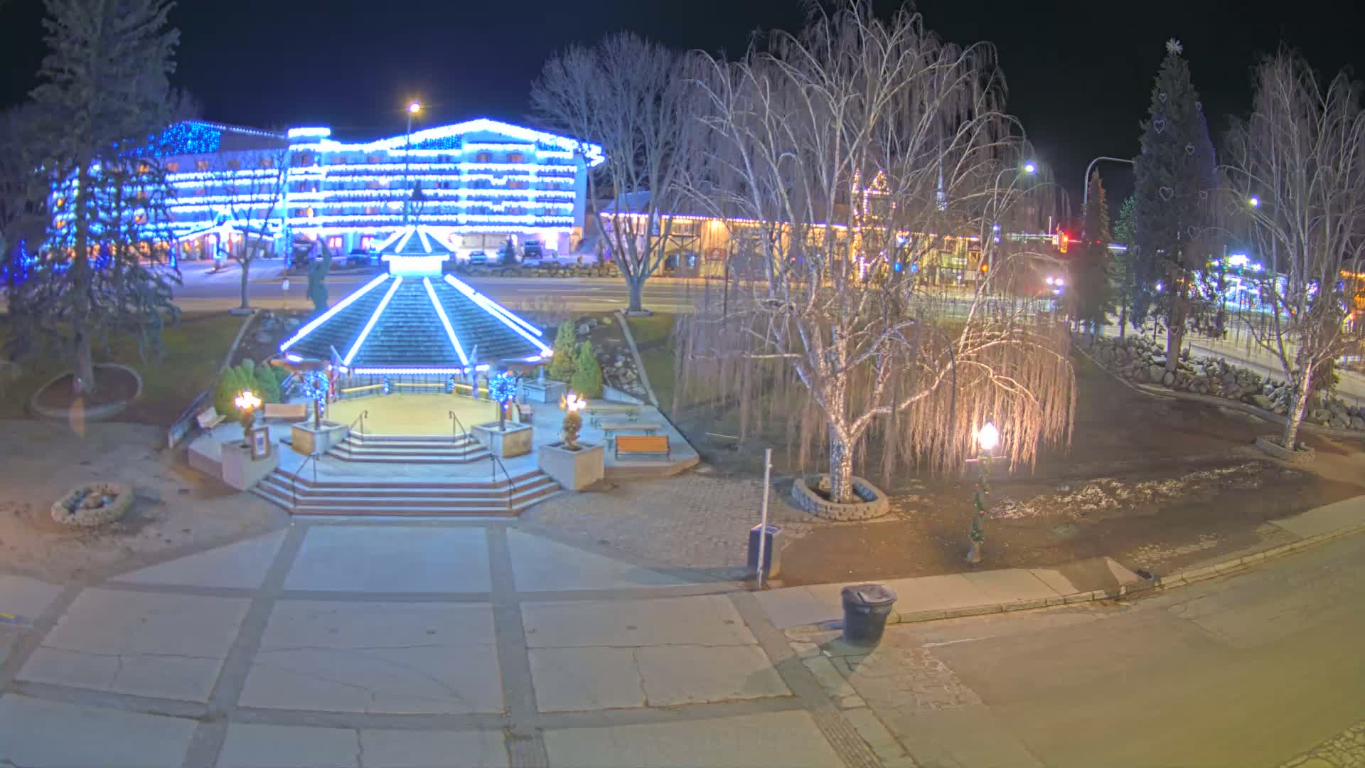 A nighttime view of a town square featuring a gazebo with blue lights, a large building also decorated with blue lights, and several bare trees under a dark sky.