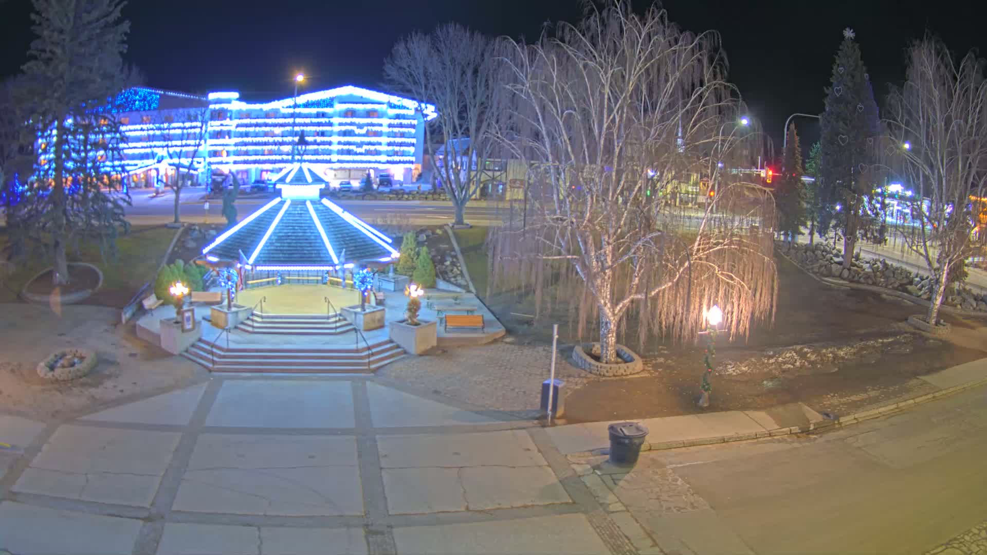 A nighttime view of a town square featuring a lighted gazebo, bare trees, a large building adorned with Christmas lights, and a decorated Christmas tree.