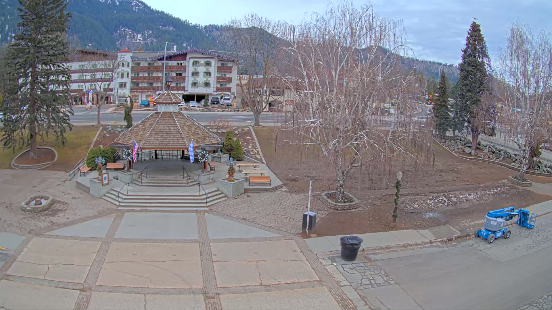 A gazebo sits in a town square, next to a mostly bare tree, under an overcast sky, with a multi-story building and mountains in the background.