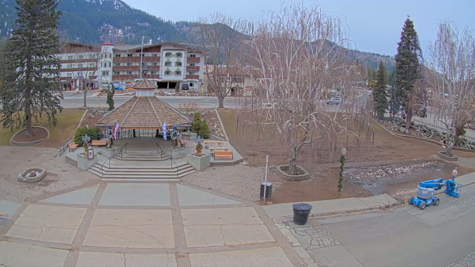 An elevated view of a town square with a gazebo, benches, bare trees, and a hotel in the background on a cloudy day.