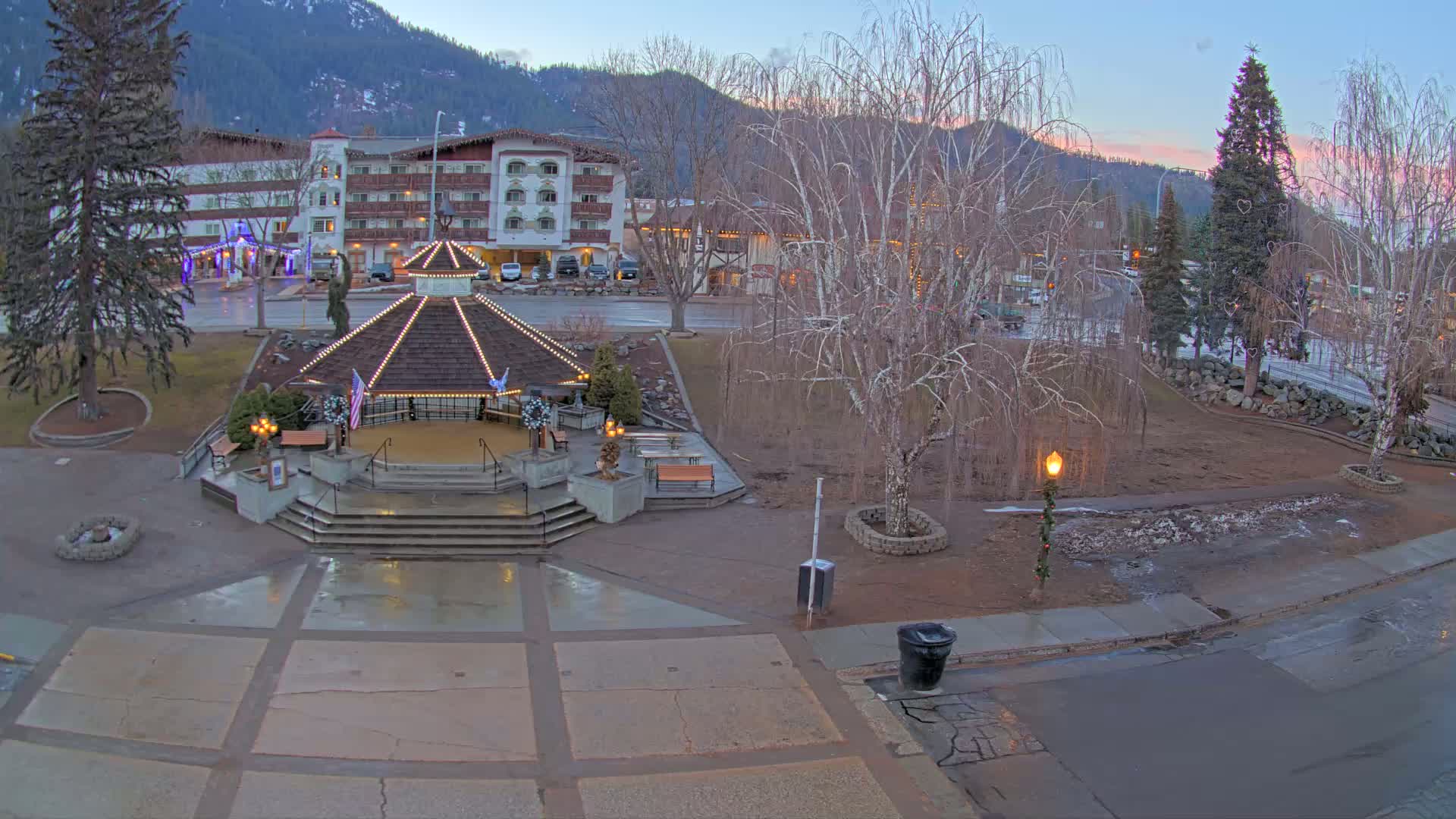 A lighted gazebo sits in a town square at dusk, with a hotel and mountains in the background, under a partly cloudy sky.