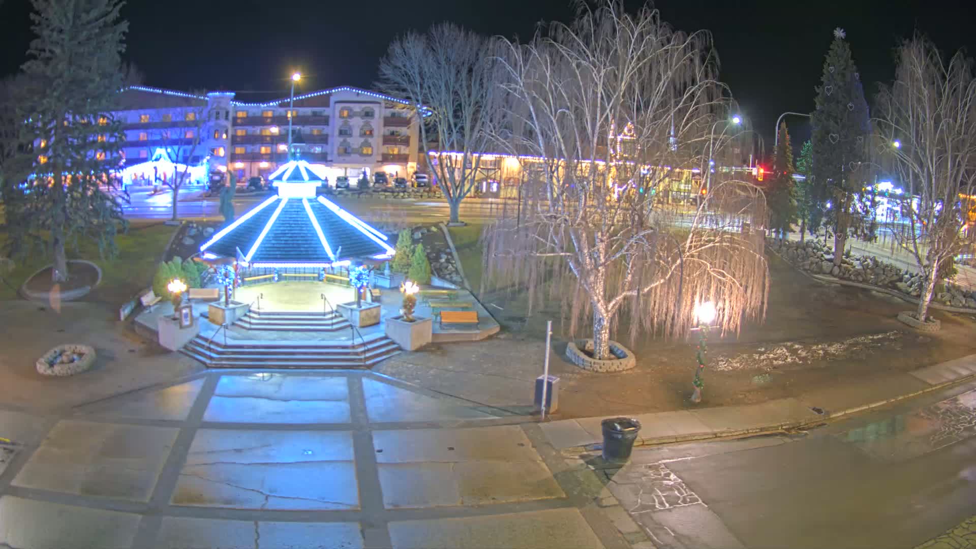 A brightly lit gazebo stands in a town square at night, surrounded by bare trees, some with Christmas lights, and a building with holiday decorations in the background, under partly cloudy skies.