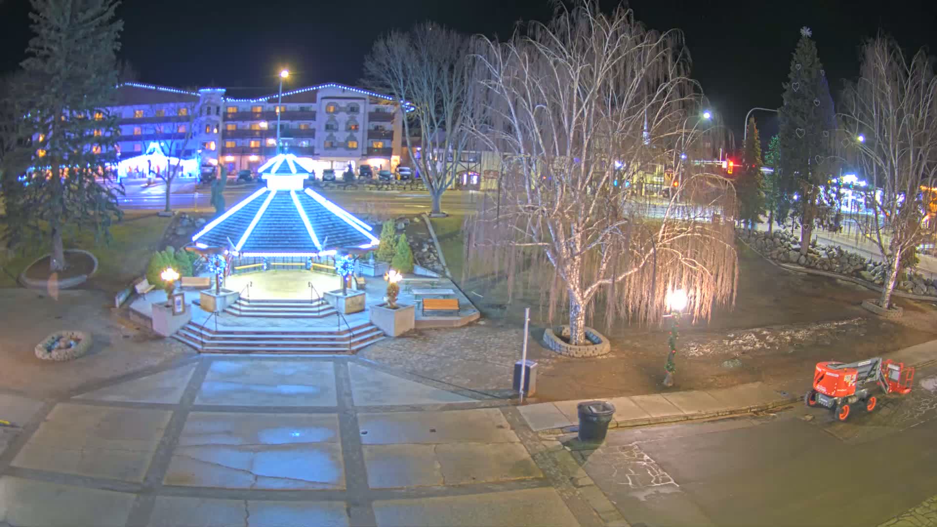 A nighttime view of a town square featuring a gazebo illuminated with blue lights, bare trees, and a small piece of construction equipment parked nearby.