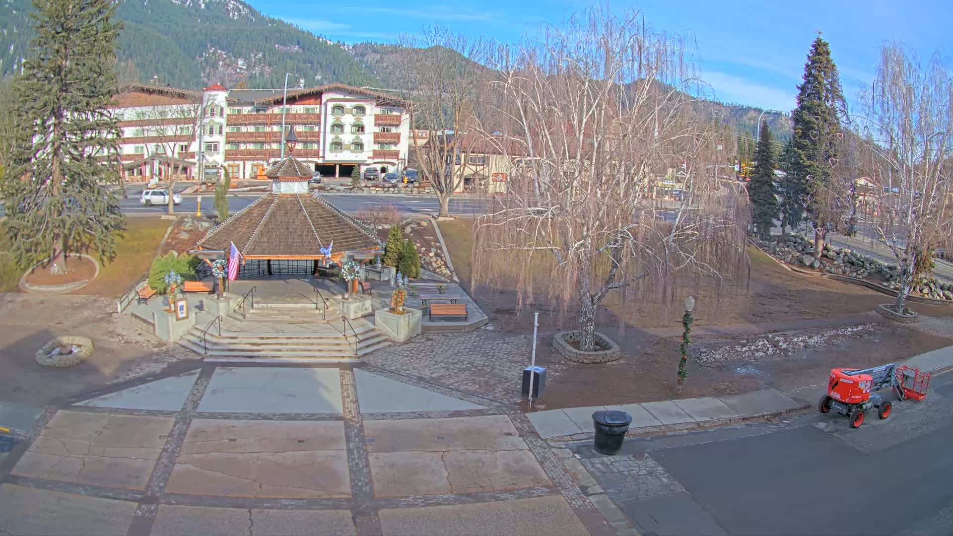 A gazebo with a paved plaza sits in front of a multi-story building on a sunny winter day, with bare trees and a small piece of construction equipment nearby.