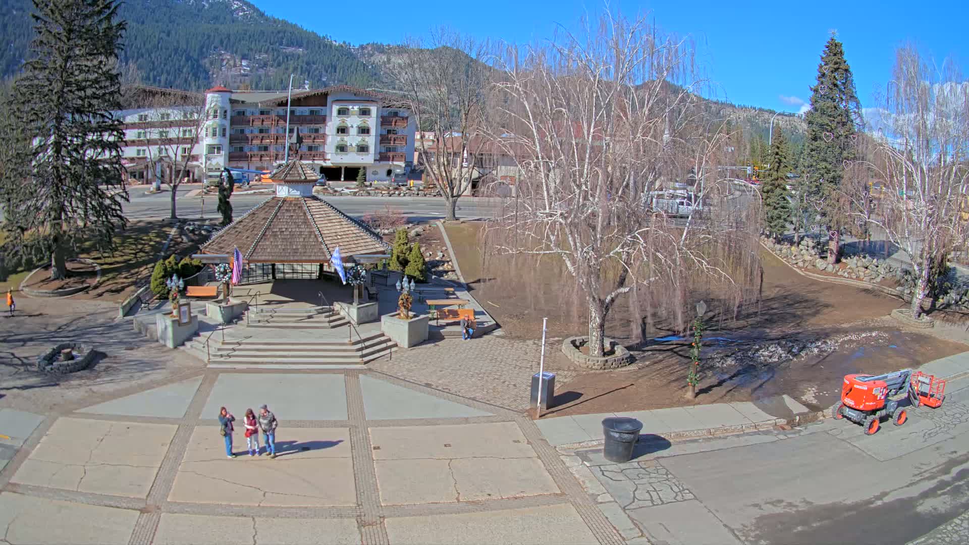 A paved town square with a gazebo, three people standing near it, and a large building in the background, all under a sunny sky.