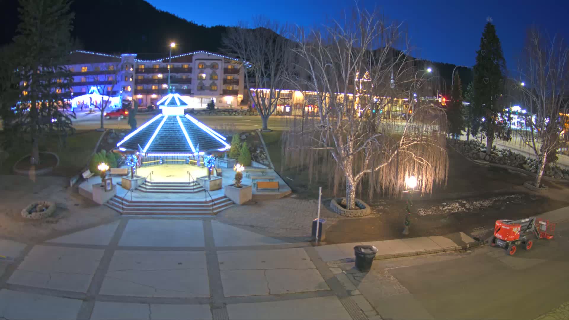 A nighttime view of a town square with a gazebo illuminated in blue lights, surrounded by bare trees and a multi-story building in the background under a clear, dark sky.