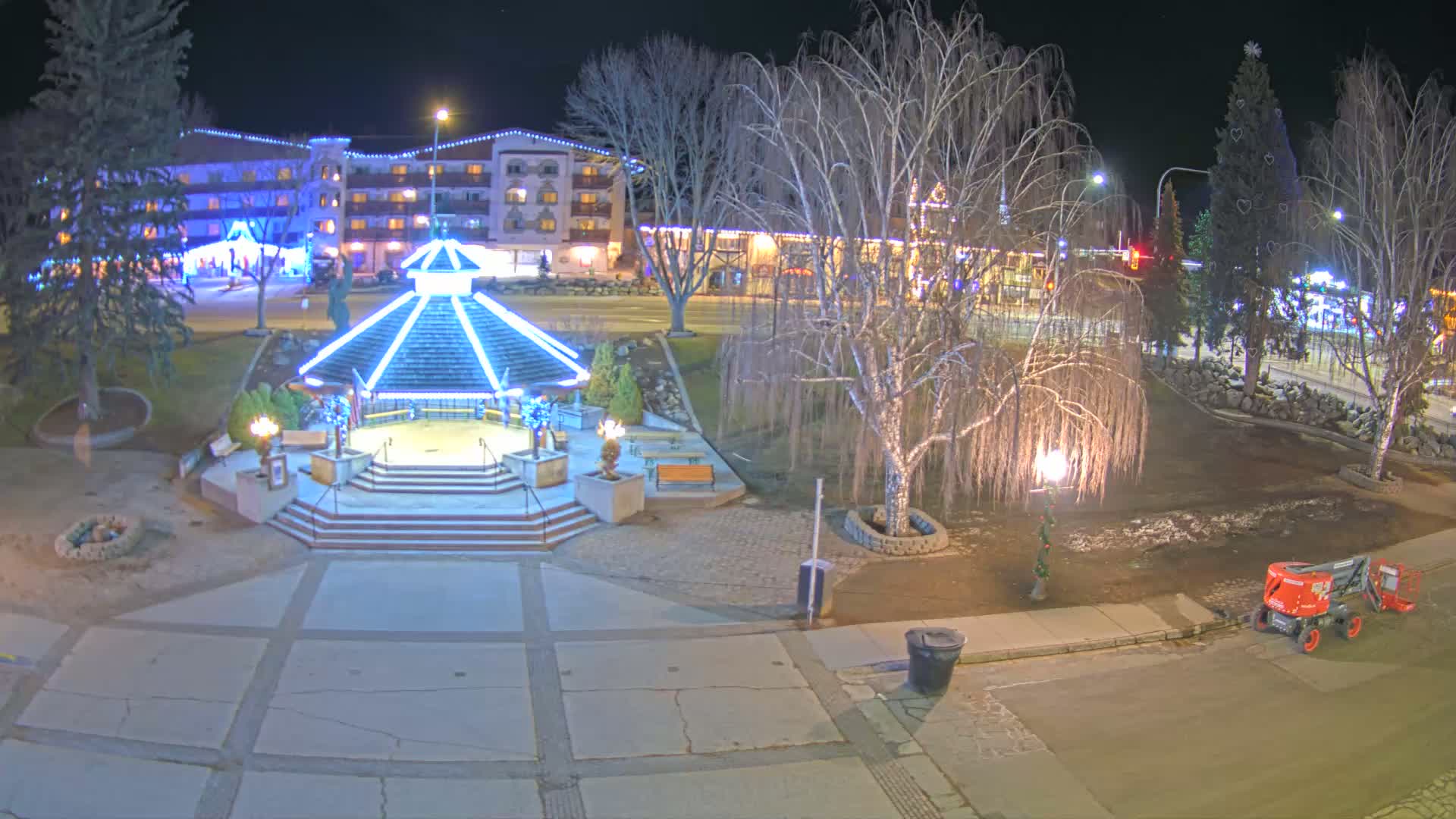 A nighttime view of a town square with a gazebo illuminated in blue lights, bare trees, and a building in the background, under a clear night sky.