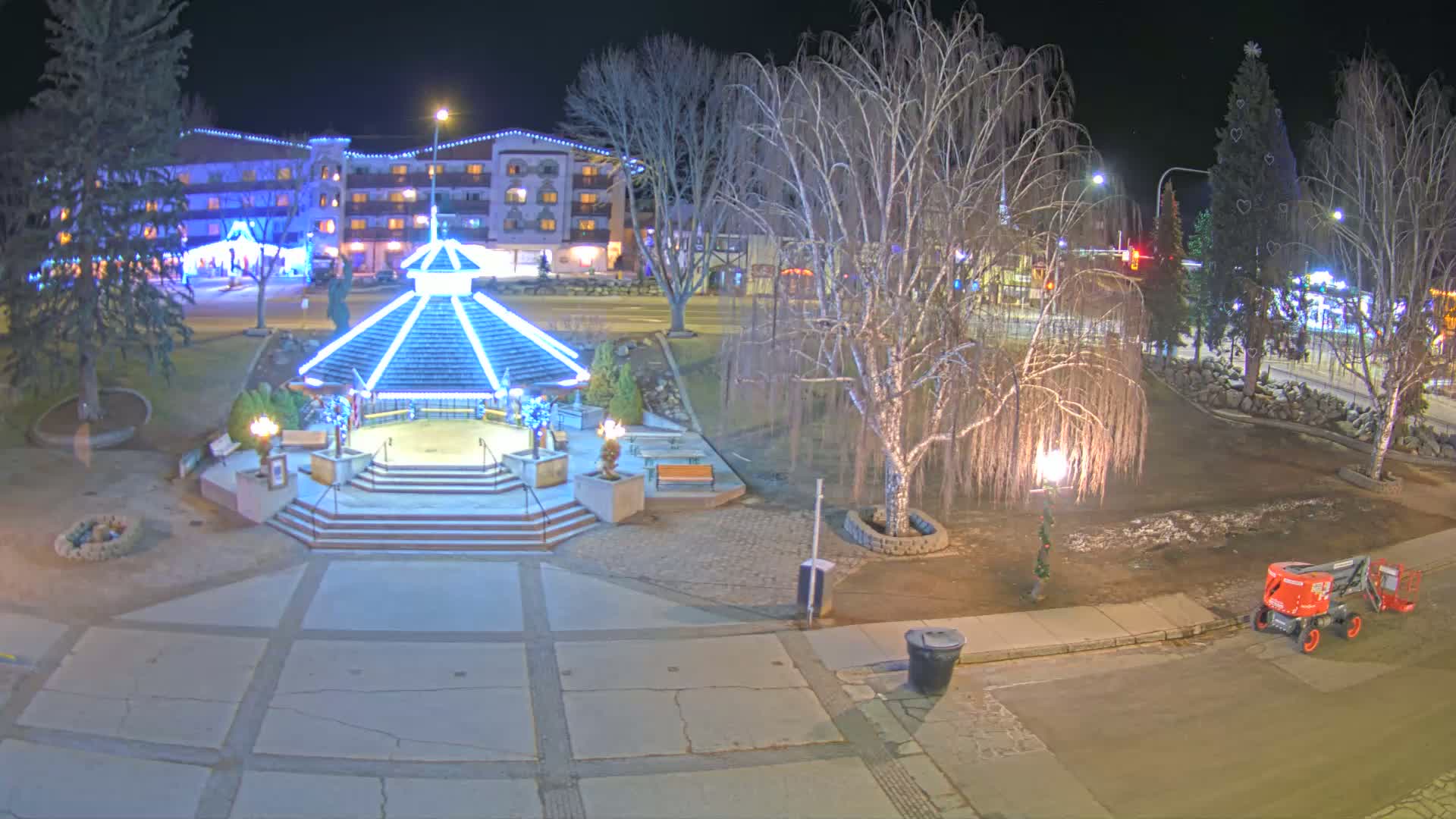 A nighttime view of a town square featuring a gazebo with blue lights, bare trees, a decorated Christmas tree, and a small piece of construction equipment.