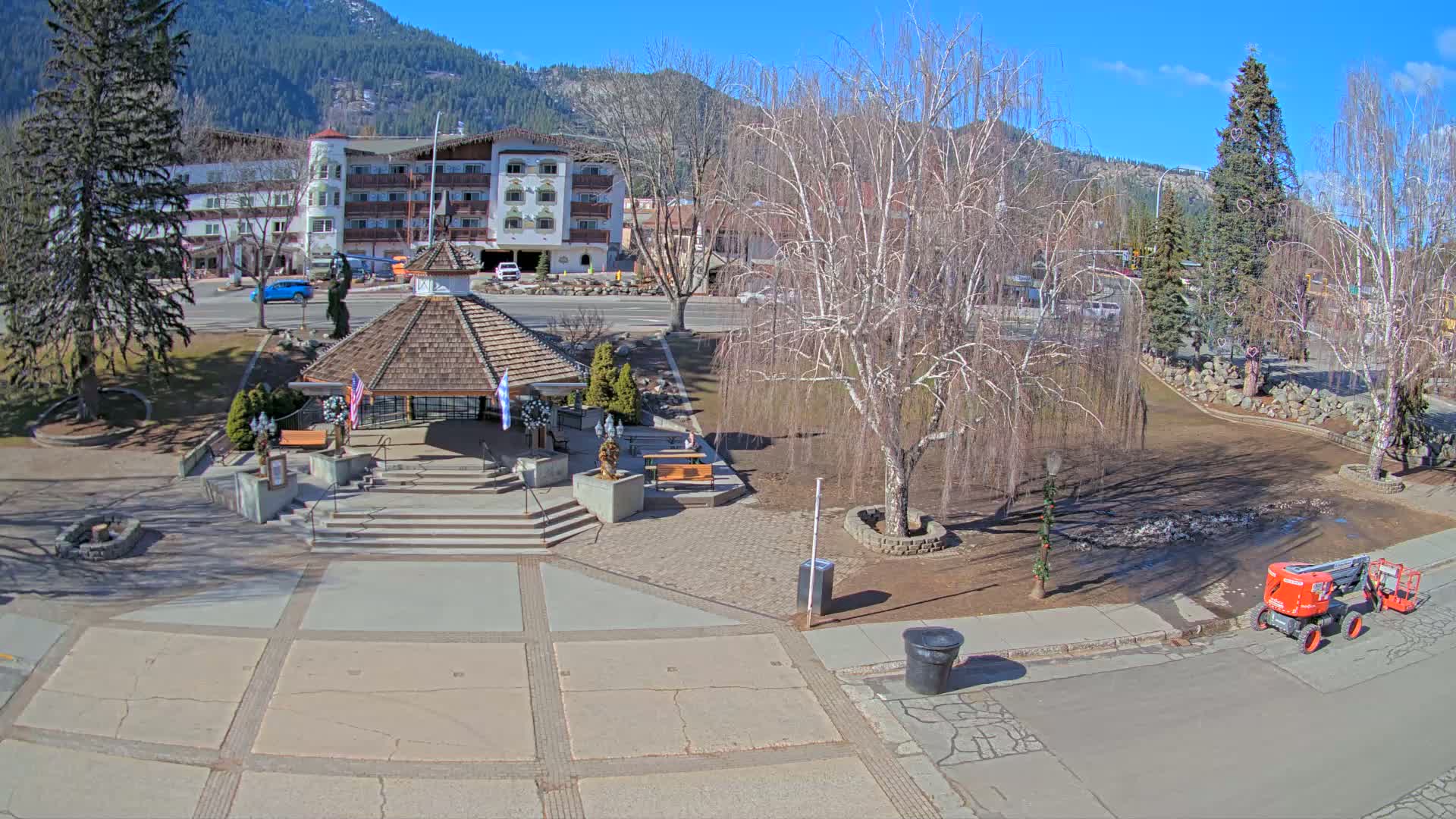 A sunny day reveals a town square with a gazebo, bare trees, a multi-story building in the background, and a small piece of construction equipment in the foreground.