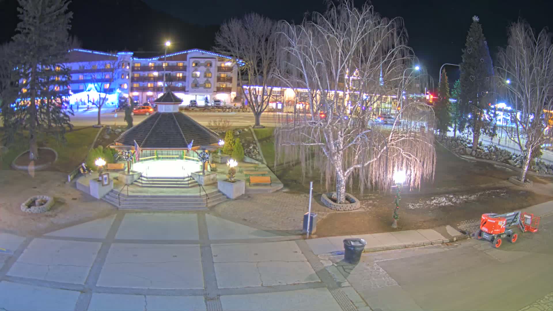 A nighttime view of a town square with a gazebo, a lit-up hotel in the background, and bare trees under partly cloudy skies.