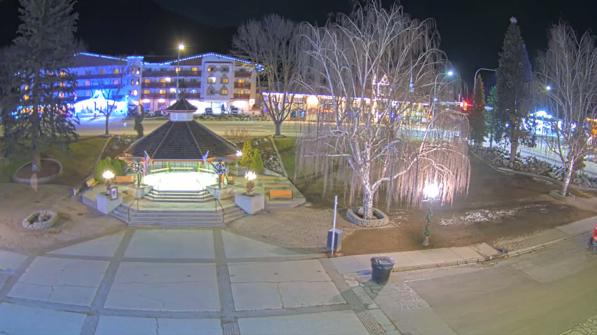 A nighttime view of a town square with a gazebo, bare trees, and a multi-story building decorated with lights.