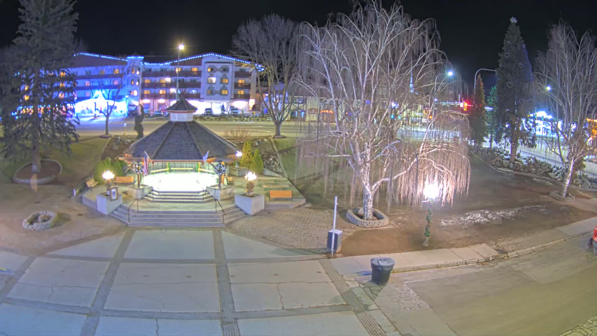 A nighttime view of a town square with a gazebo, bare trees, and a building decorated with lights, with patches of snow on the ground.