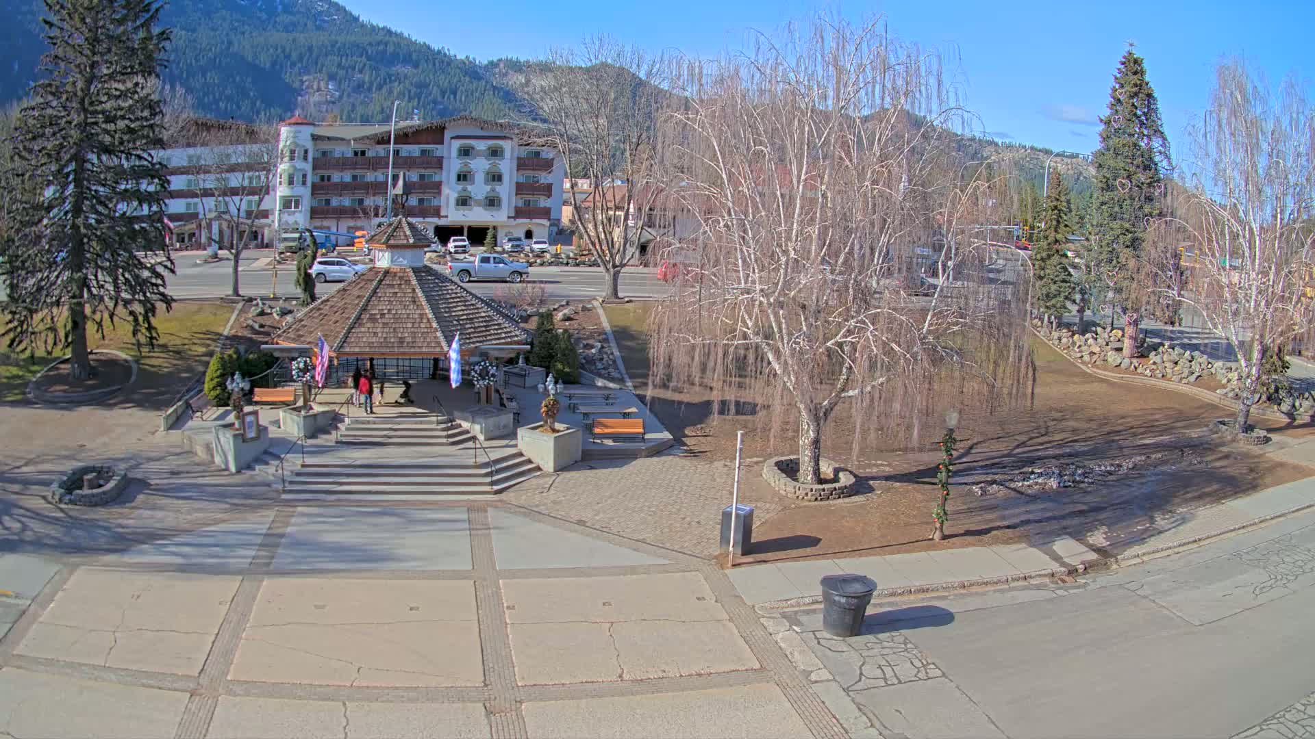 A gazebo sits in a town square on a sunny day, with a multi-story building visible in the background and bare trees surrounding the plaza.