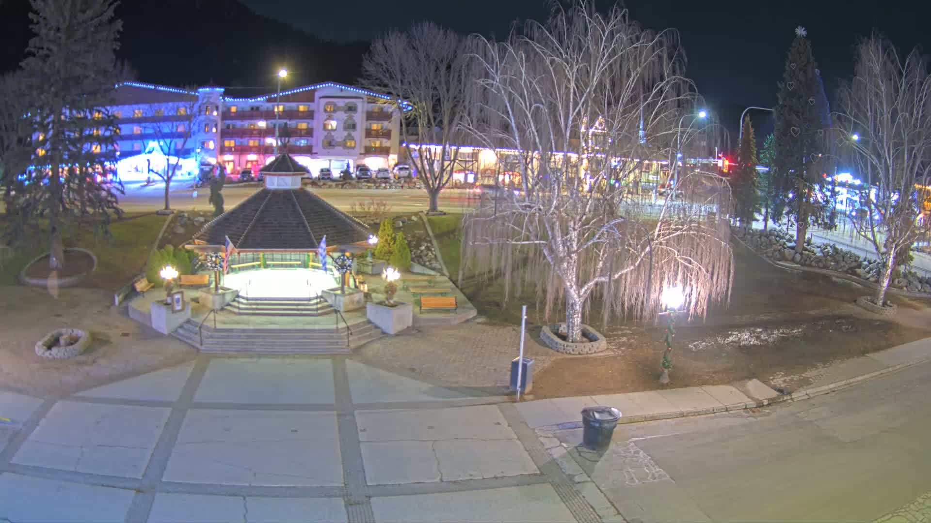 A nighttime view of a town square with a gazebo, bare trees, a brightly lit hotel, and a decorated Christmas tree.