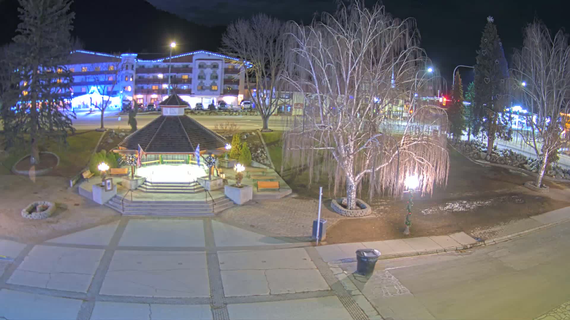A nighttime view of a town square with a gazebo, trees, and a hotel in the background, under partly cloudy conditions.