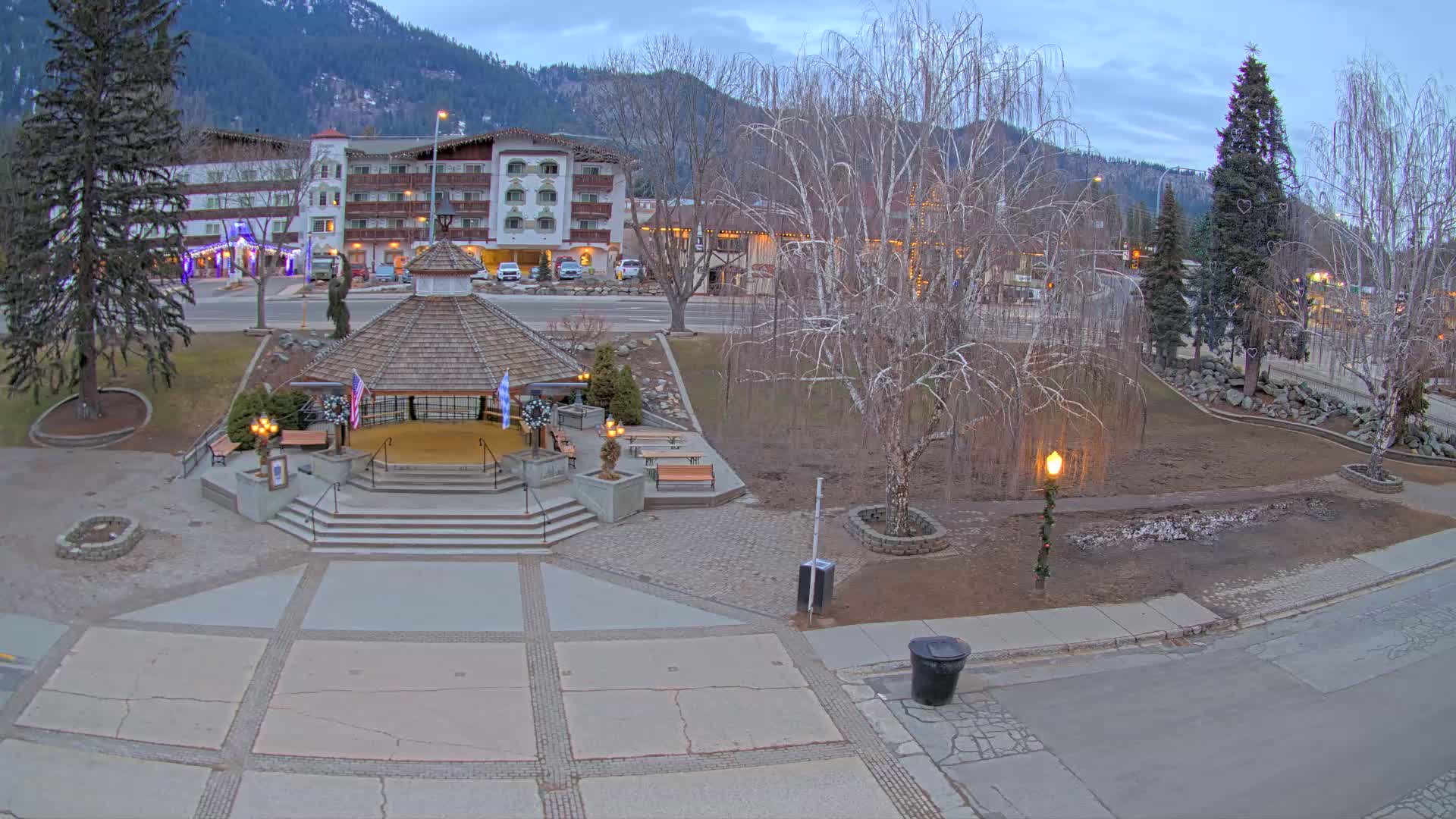 A gazebo sits in a town square at dusk, surrounded by bare trees and a multi-story building in the background under a cloudy sky.